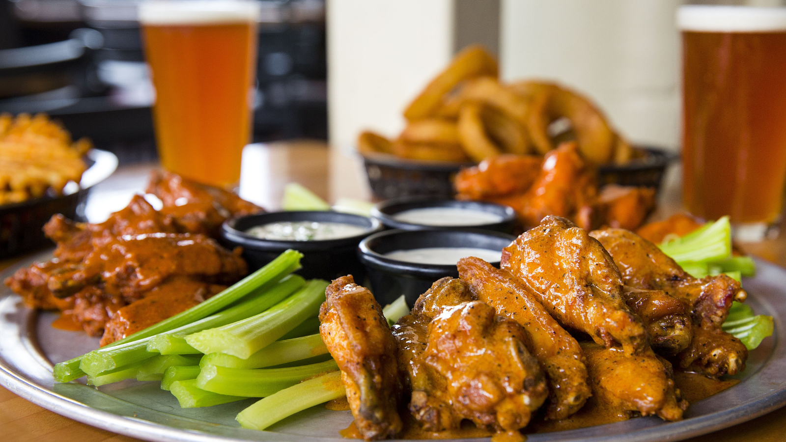 Close up of plate of chicken wings, sauces and cut celery. In the back is a glass of beer and bowl of onion rings.